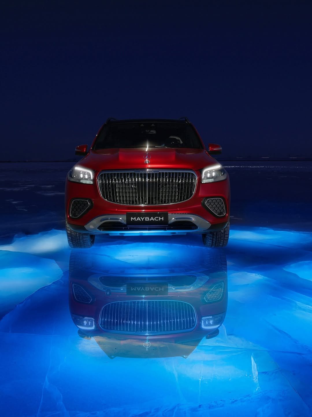 Front view of a red Mercedes-Maybach GLS car on reflective blue surface. The car's headlights are on, and the background is a dark blue sky. The word "MAYBACH" is visible on the car's front.