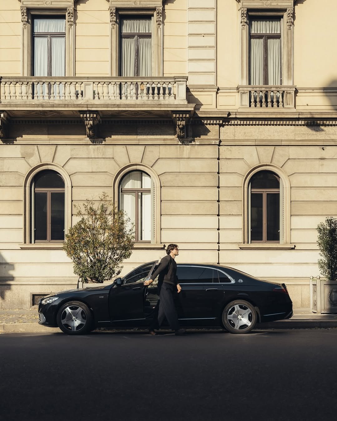 A man exits a dark Mercedes-Maybach S-Class parked alongside a light-colored building with arched windows. The man wears dark pants and a sweater. The car's door is open. Sunlight casts shadows on the facade.