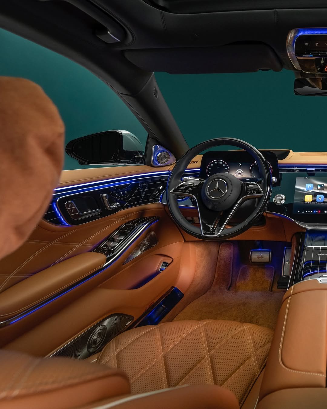 The interior of a luxury Mercedes-Maybach S-Class with light brown leather seats and trim, ambient blue lighting, and a digital display showing parking assist, FM/DAB radio, and Bluetooth player options, plus a speedometer reading 0 km/h. The steering wheel features a Maybach logo.