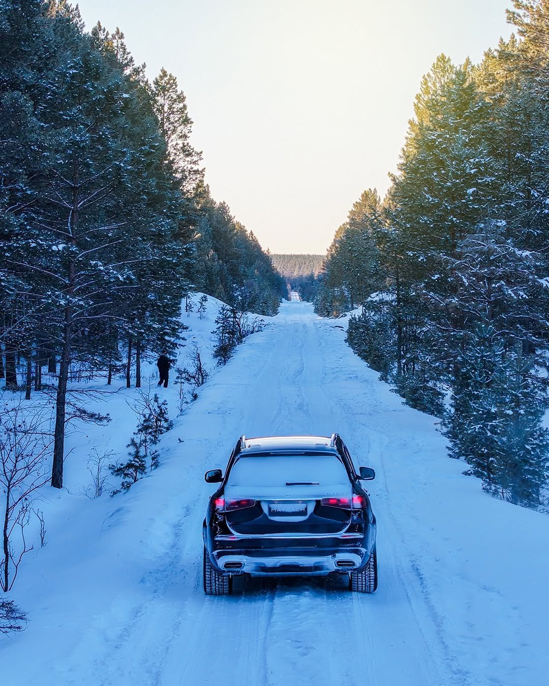 A Mercedes-Maybach GLS drives on a snowy road between trees, heading away from the viewer. There is a person walking beside the road. The car has snow on its roof and has a license plate.
