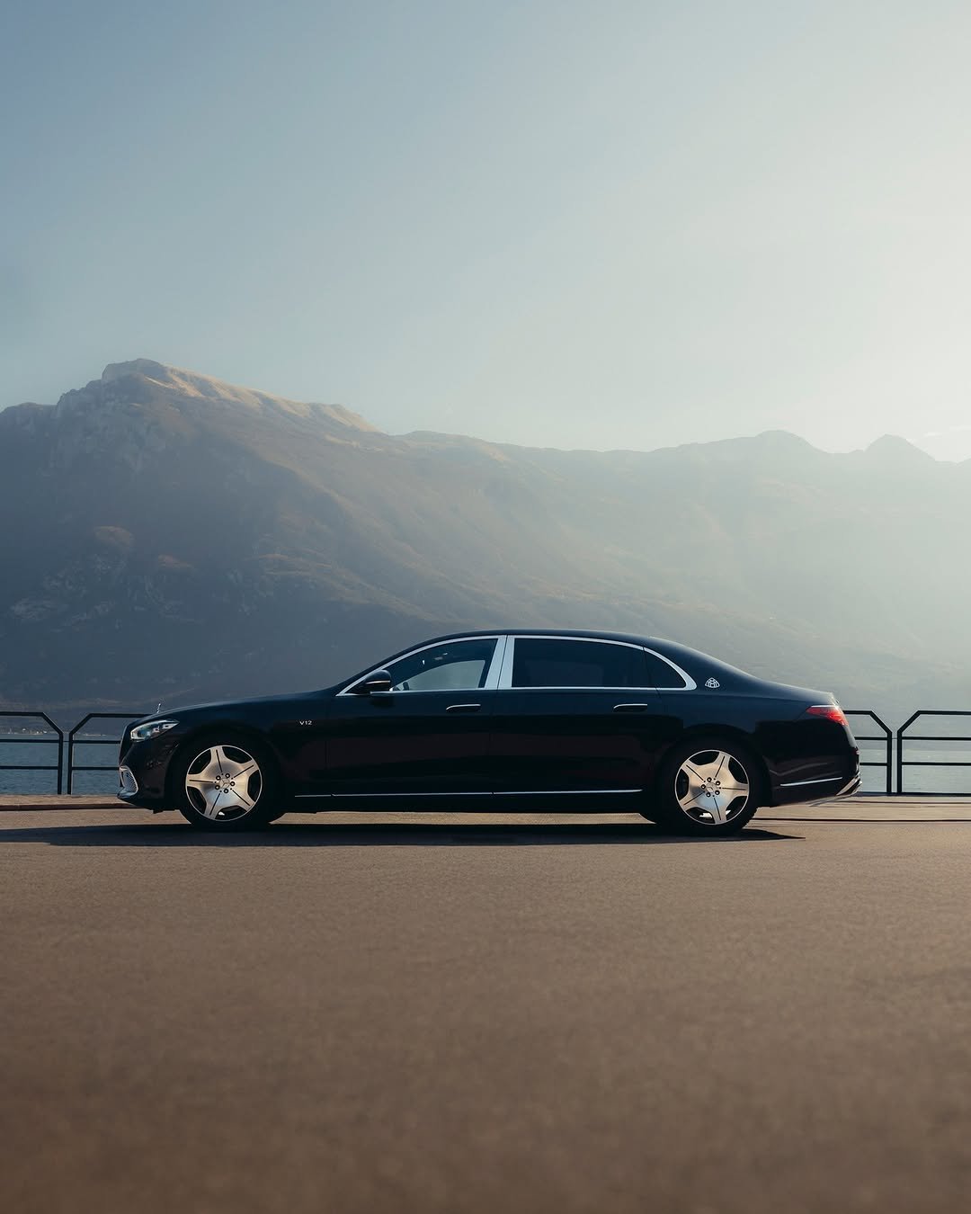 A dark Mercedes-Maybach S-Class is parked on a paved surface with mountains in the background. The car's features include shiny rims and a sunroof. A body of water and a railing are subtly visible behind the car.