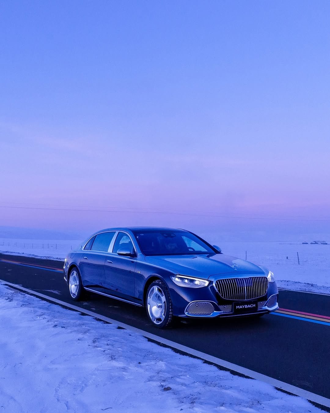 A dark blue Maybach stands on a road with snow on the sides under a twilight sky. The car faces the camera, displaying its front grill and headlights. The brand 'MAYBACH' is visible on the car's front.