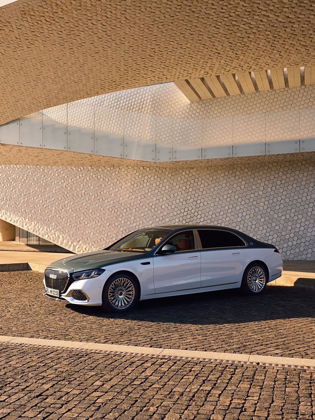 A white and green Mercedes-Maybach S-Class, with a license plate "S MM 2230", is parked on a brick road. The car is parked in front of a large building that has a light tan pattern of hexagon-shaped tiles.