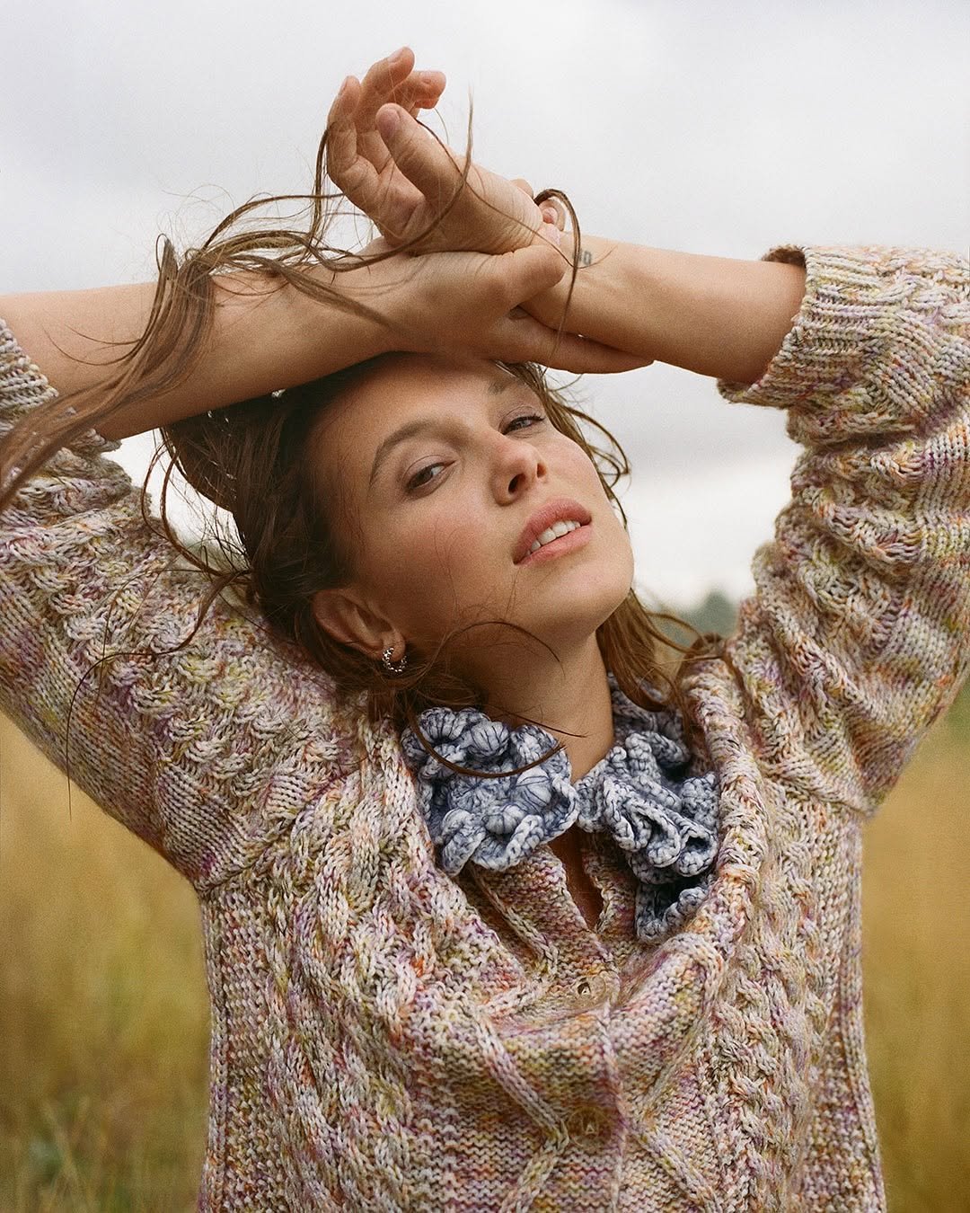 Image one shows Millie Bobby Brown in close up in a field, wearing a beige chunky knit jumper. She has both arms raised and the backs of her hands to her forehead, and she is looking into the camera.
