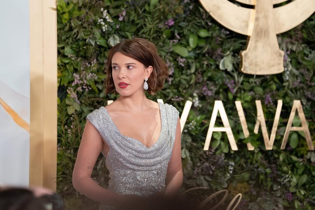 Photo by Millie Bobby Brown on January 20, 2026. May be an image of one or more people, makeup, dress, carpet, gown and text that says 'AWA'.
