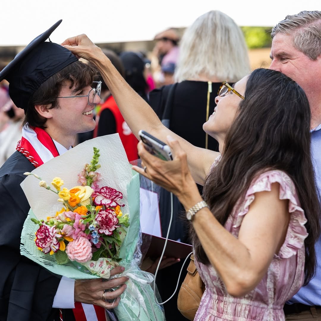 A person in a graduation cap and gown receives a bouquet of colorful flowers from another person, who is adjusting the graduate's cap.