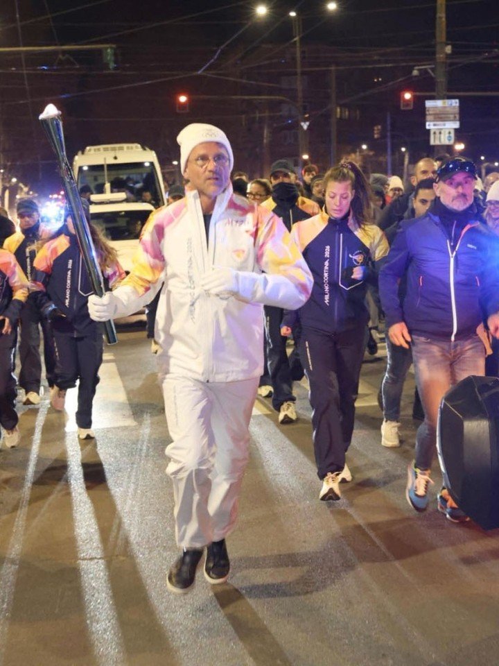 Carlo Ratti in a white outfit carrying an Olympic torch, leading a group of individuals in matching jackets down a city street. Emergency vehicle lights flash in the background as they proceed in a night setting.