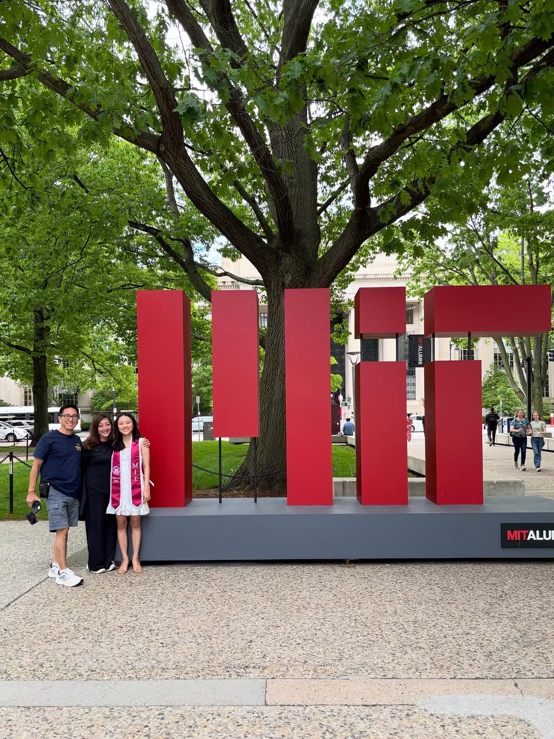 A family of three smiling in front of the red MIT sign on campus.