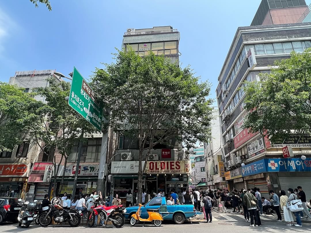 Photo by 올디스 타코 on July 02, 2025. May be an image of 9 people, scooter, street, skyscraper, buildings and text.