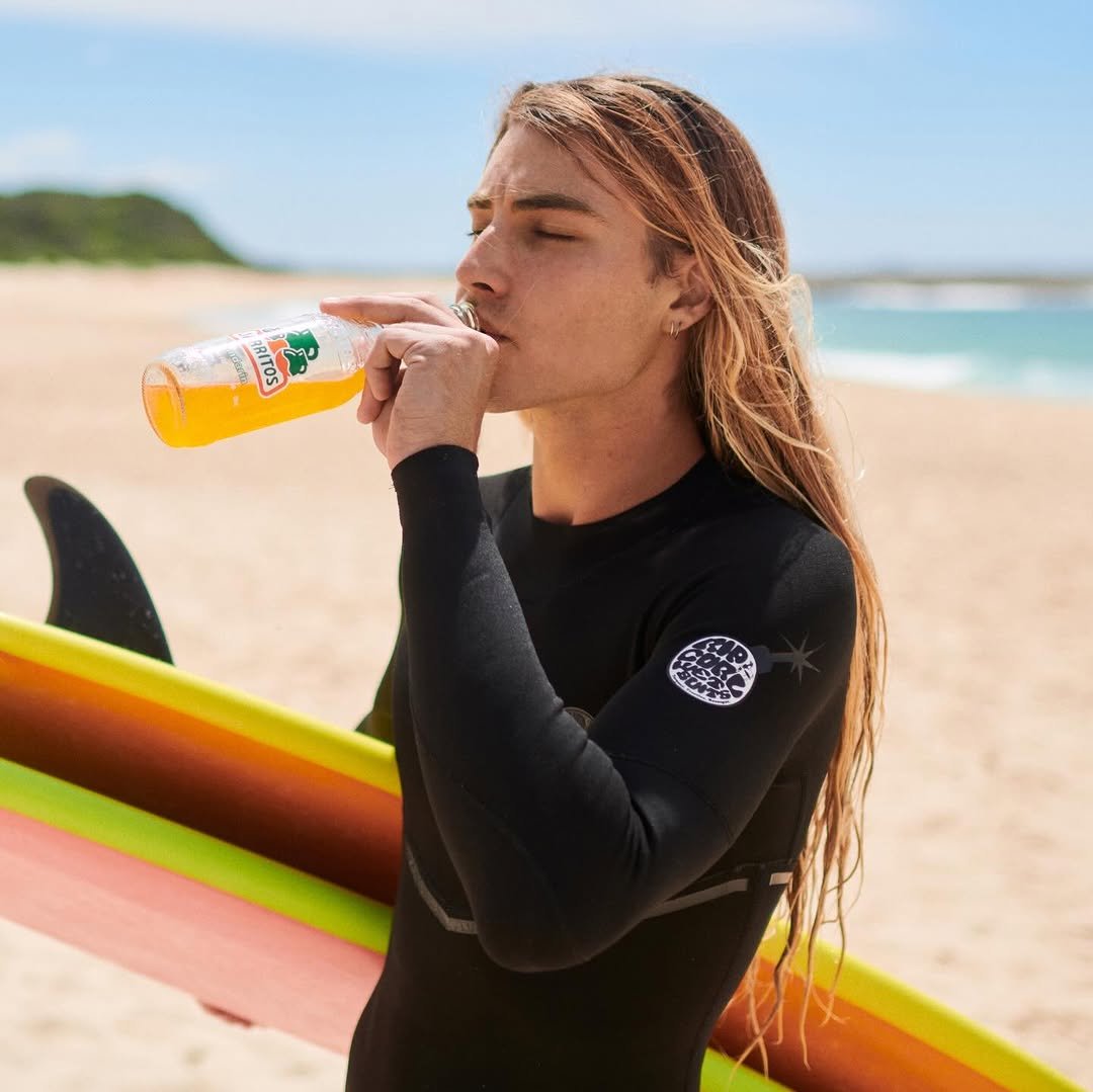 Photo shared by Oscar Langburne on December 19, 2025 tagging @jarritos, and @jarritosau. May be an image of surfboard, juice, wetsuit, bottle, water bottle, water, beach and text that says 'S RITOS'.