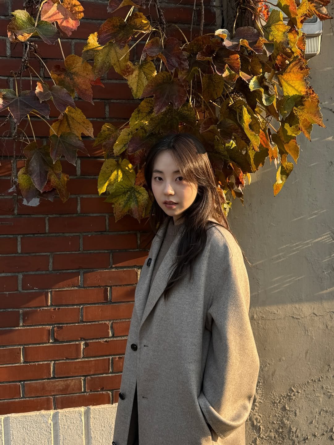Photo by 안소희 on November 24, 2025. May be an image of one or more people, hair, duffle coat, overcoat, goosedown coat, turtleneck, parka, brick wall and text.