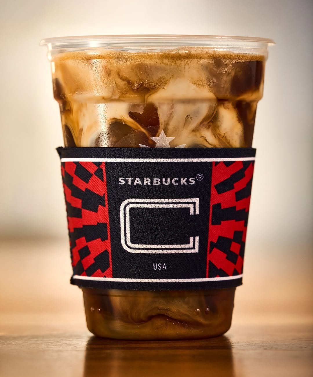 Iced coffee drink in a Starbucks cup with milk swirling through dark coffee, wrapped in a red geometric sleeve that reads “Starbucks USA