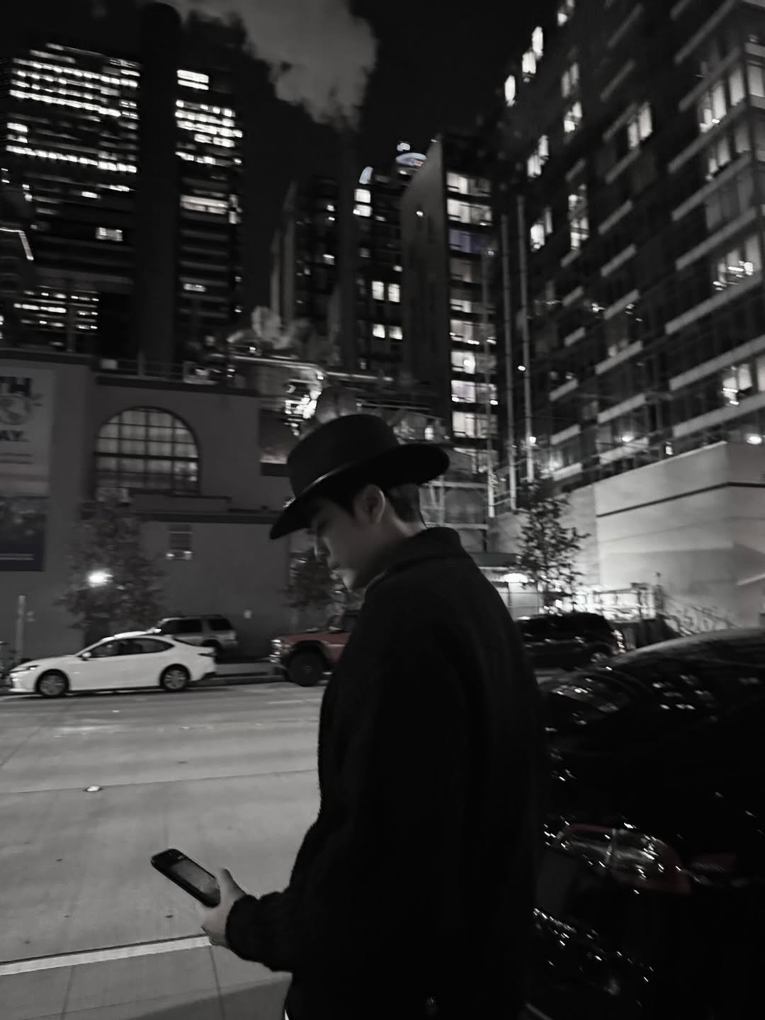 Photo by 김수겸 on January 11, 2026. May be a black-and-white image of smoking, hat, phone, overcoat, cigarette, bowler hat, street, skyscraper, night and text.