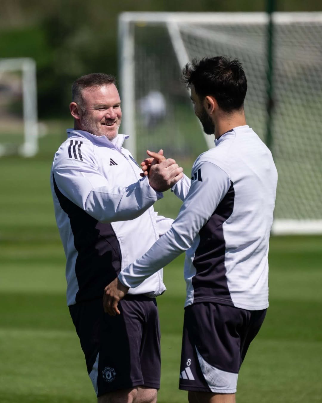 Wayne Rooney and Bruno Fernandes in training gear shake hands on a green field with goalposts in the background.