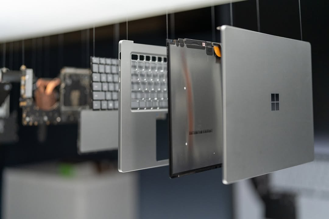 A series of disassembled laptop components hanging in mid-air, including the keyboard, motherboard, and casing of a Microsoft laptop. The arrangement showcases the intricate details of each part against a blurred background.