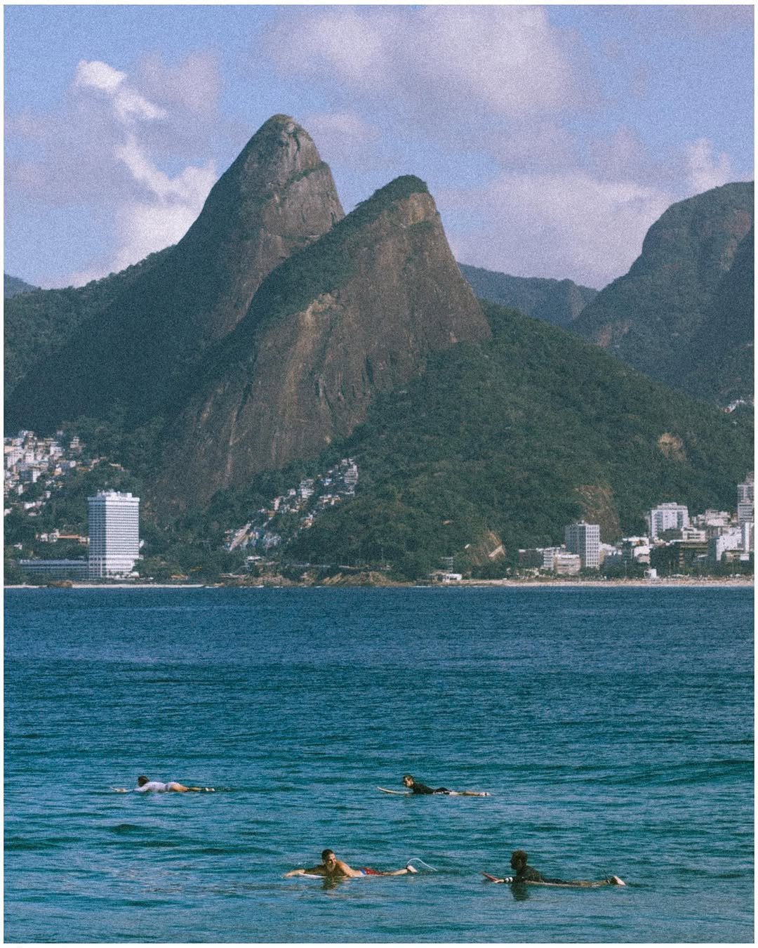 A mountainous coastal landscape with tall, rocky peaks overlooking an ocean, with several people swimming in the water in the foreground.