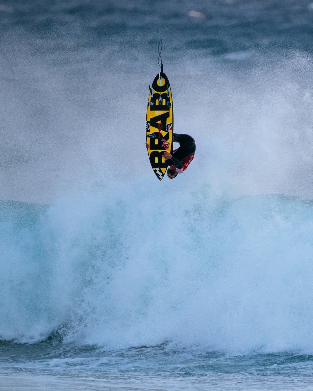 A person performing an aerial stunt on a yellow and black surfboard while riding a large wave.