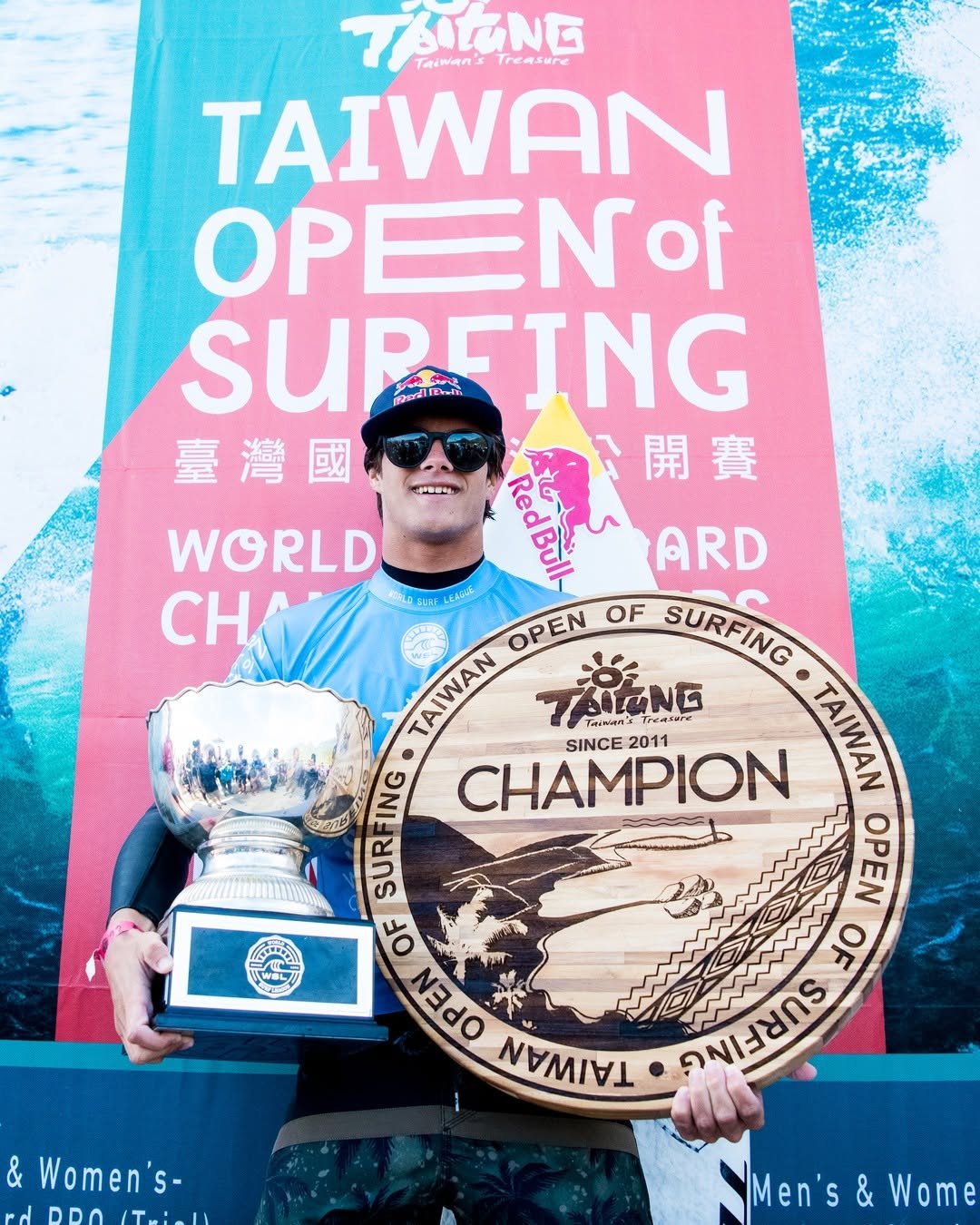 A person wearing sunglasses and a hat is holding a trophy and a large circular award while standing in front of a banner announcing the "Taiwan Open of Surfing" event.