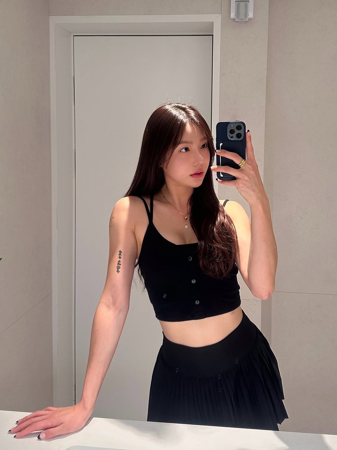 거울 앞에서 블랙 버튼 탑과 플리츠 스커트를 입고 셀카를 찍는 여성