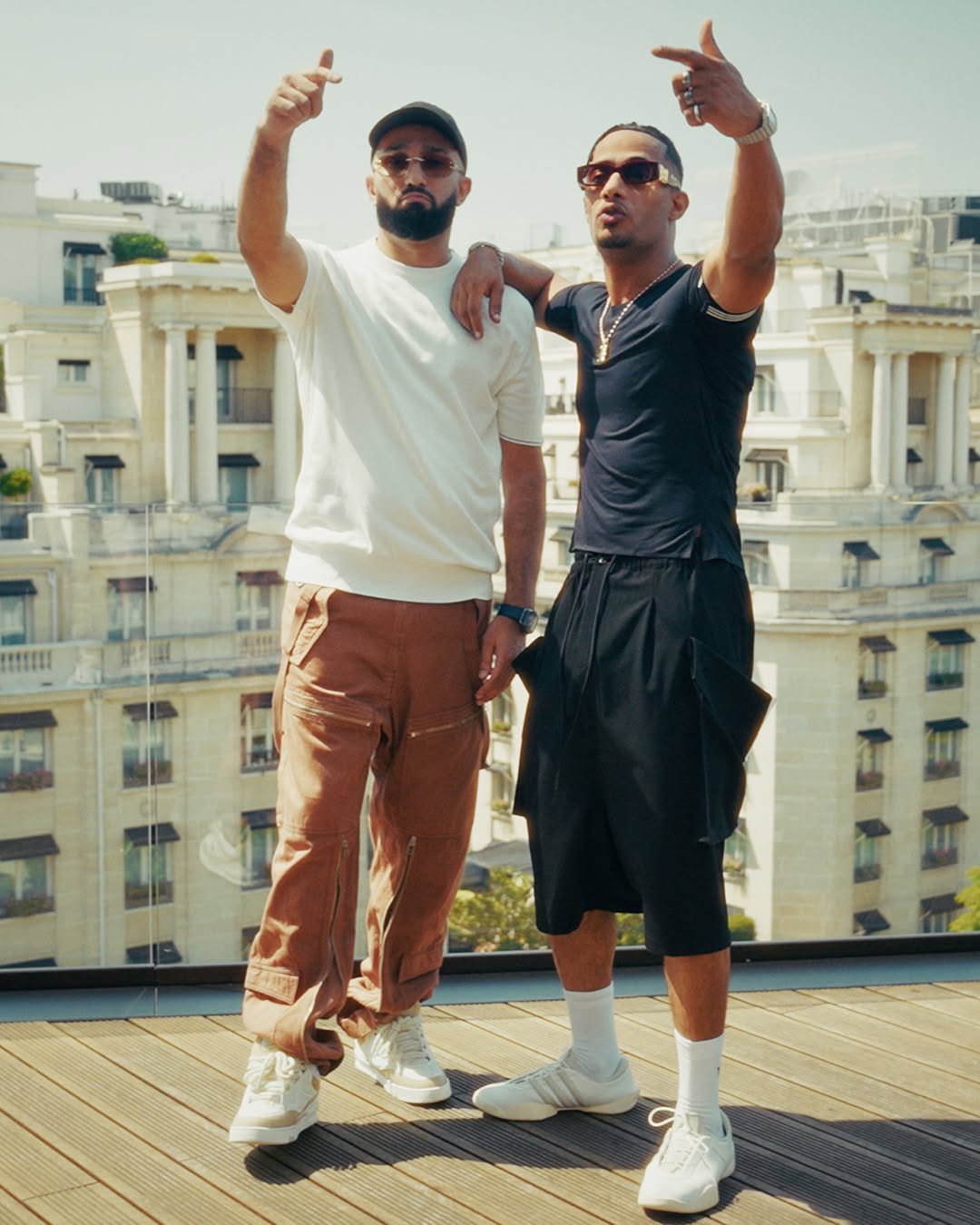 MR1 & Niro stand together on a sunlit wooden balcony posing with their fingers pointed up, wearing casual streetwear including a white t-shirt with brown zippered cargo pants and a black shirt with baggy black shorts against a bright backdrop of classic multi-story city buildings.
