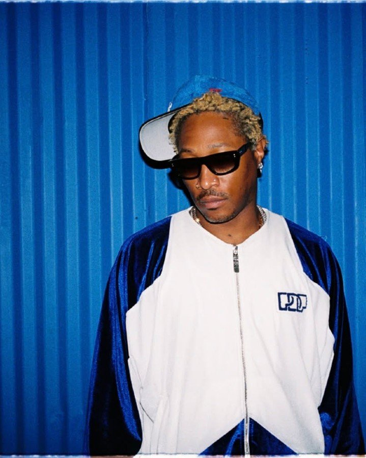 Future poses in front of a bright blue corrugated metal wall, looking slightly downward. He wears dark sunglasses, a tilted blue baseball cap over short bleached hair, and a zippered white track jacket with royal blue sleeves.
