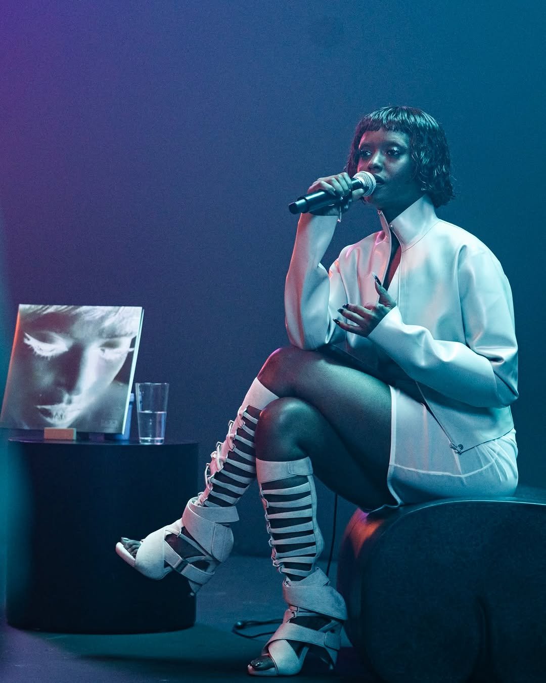 Kelela sits on a curved stage prop under dramatic blue and purple lighting, speaking into a microphone. She is dressed in a white jacket, a white skirt, and white knee-high strappy gladiator sandals. Beside her on a small black table rests a glass of water and a record sleeve featuring a close-up portrait.