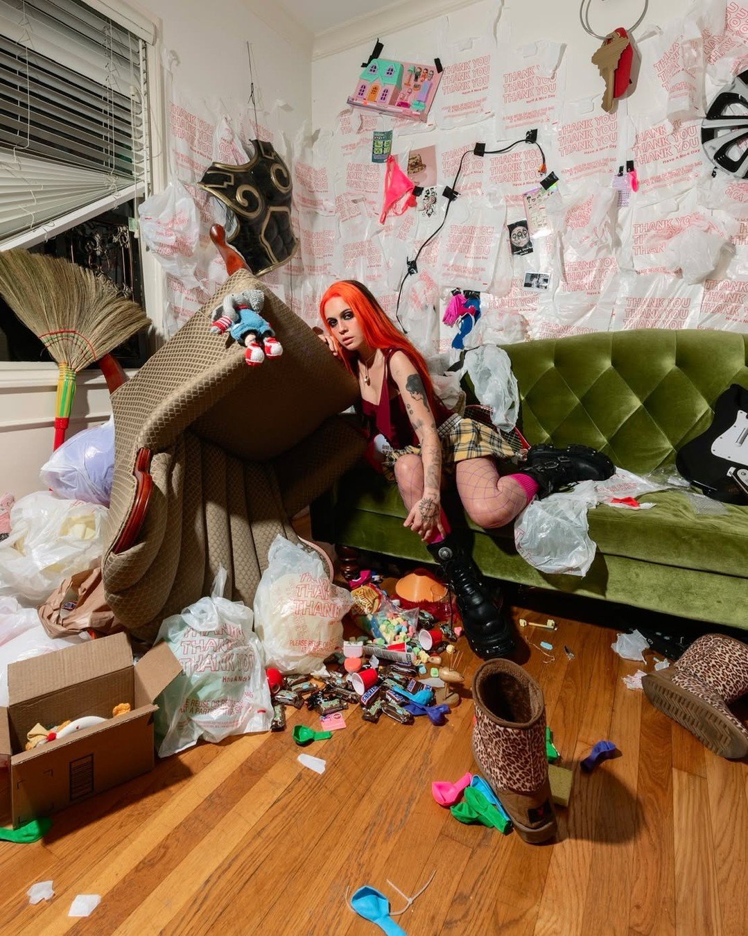 Image of Bea Miller with bright orange and black hair sitting on a green velvet couch in a chaotic, highly cluttered room. She is wearing a maroon tank top, yellow plaid mini skirt, fishnet tights, and chunky black platform boots. The walls behind her are completely plastered with white plastic "Thank You" grocery bags, and the floor is covered in scattered debris including candy, deflated balloons, and an overturned armchair.