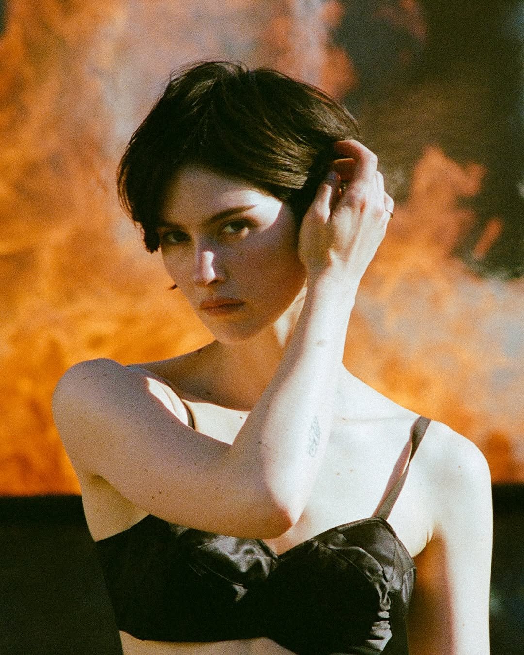 Gracie Abrams stands outdoors with a large, bright orange fire blazing in the background. She is wearing a black bralette top and has one hand resting gently near her ear, looking directly at the camera. Harsh sunlight casts defined shadows across her face and collarbone, contrasting with the dramatic flames behind her.