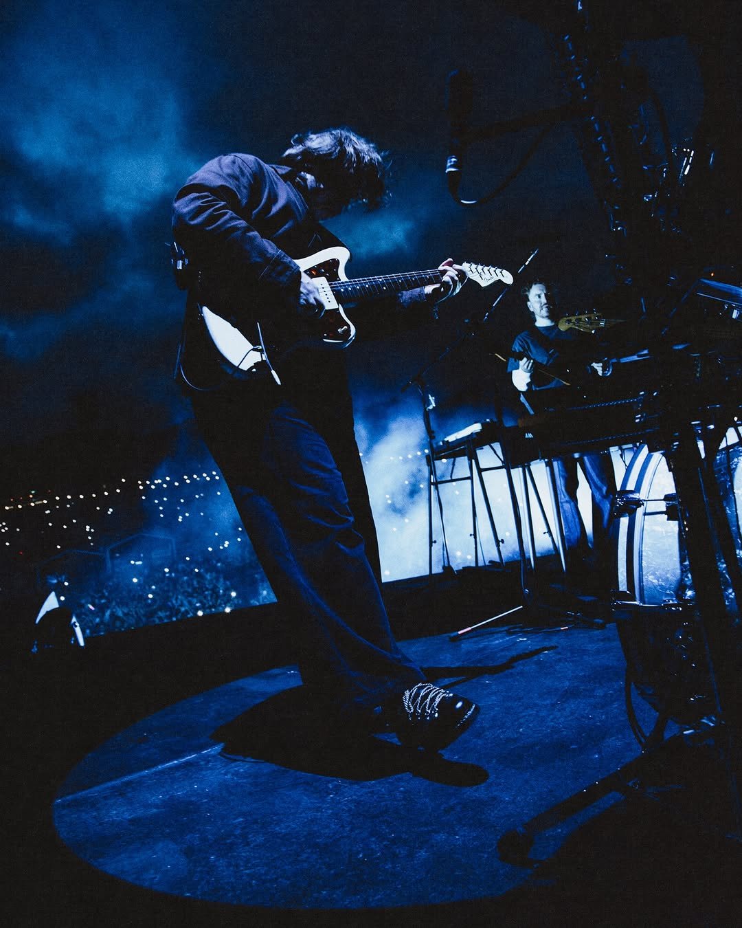A 9 image carousel showcasing Lewis Capaldi performing and rehearsing. Highlights include a dramatic blue-lit shot of him playing guitar for a massive crowd, a black-and-white close-up of him singing passionately, and vibrant stage photos where he performs in front of massive backdrops featuring a blooming pink flower and a cloudy night sky. Other photos show him rehearsing with a trumpet player under a spotlight, practicing backstage in a small room, and playing an acoustic guitar in silhouette. The final image is a poster for his BST Hyde Park concert on July 11, 2026, streaming on YouTube, featuring him singing and playing guitar under starry stage lights.